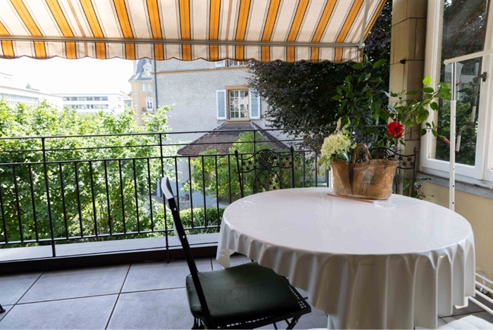 Bild vom Balkon, auf dem Balkon steht ein runder Tisch, geschmückt mit einem schönen Blumenstrass und davor steht ein schwarzer Stuhl. Die Sonnenstoren sind draussen und das Wetter ist sonnig.